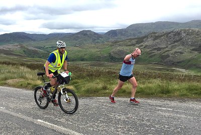 Support cyclist on road following runner through Scottish scenery