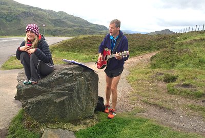 Ian Carley at roadside playing guitar and singing whilst Cyra Parks sits on rock in front